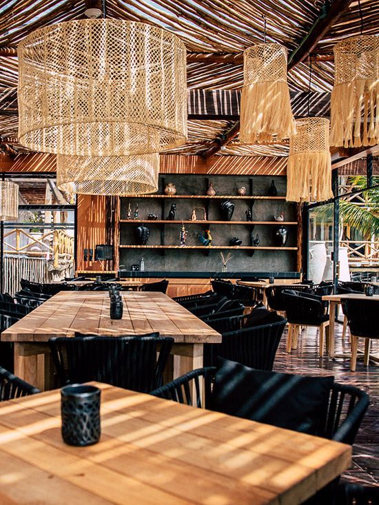 Restaurant interior with woven lamps, wooden tables, and black chairs. A shelf displays decor.