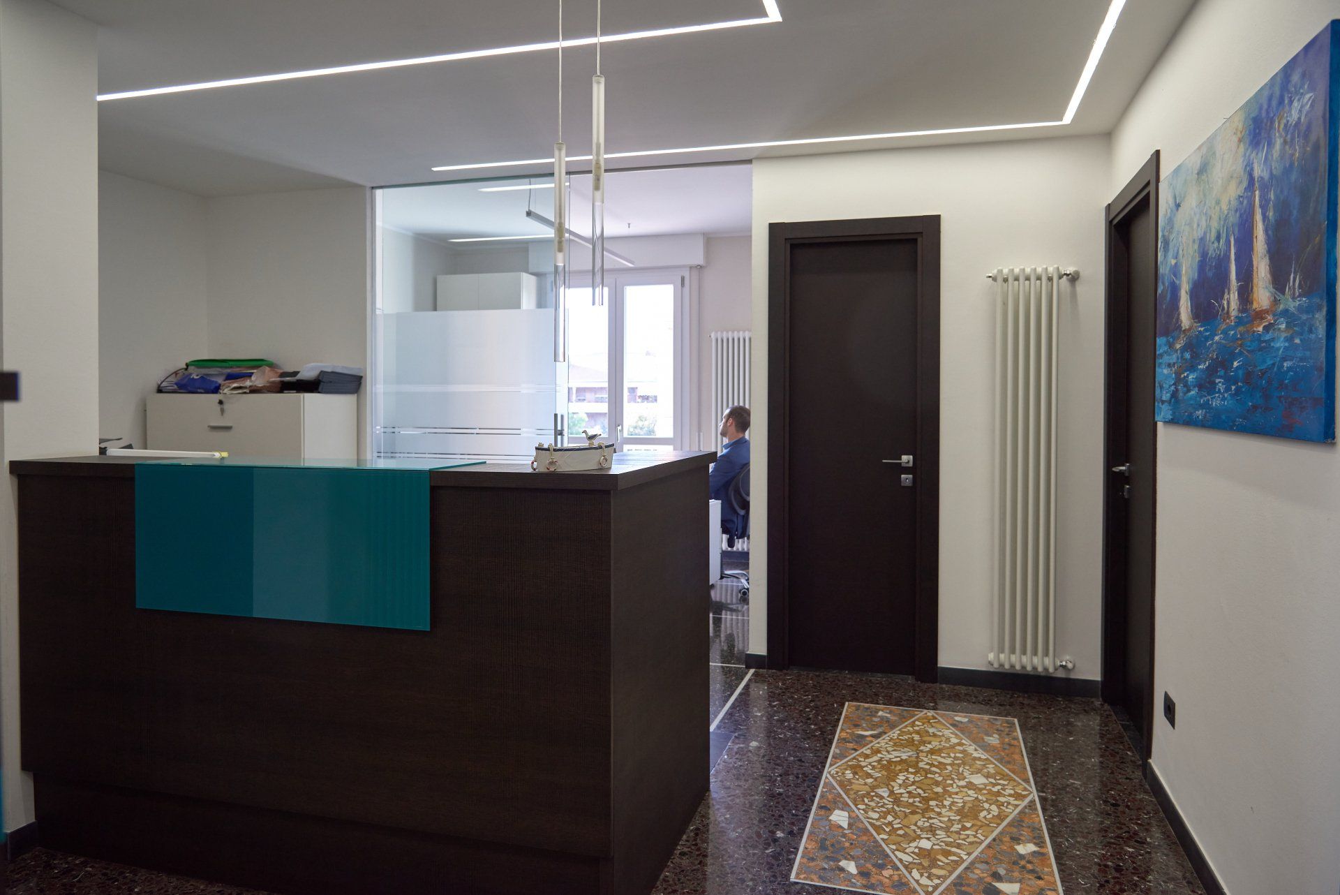 Area reception con scrivania scura, dettagli color verde acqua e sala d'attesa dietro vetro smerigliato.