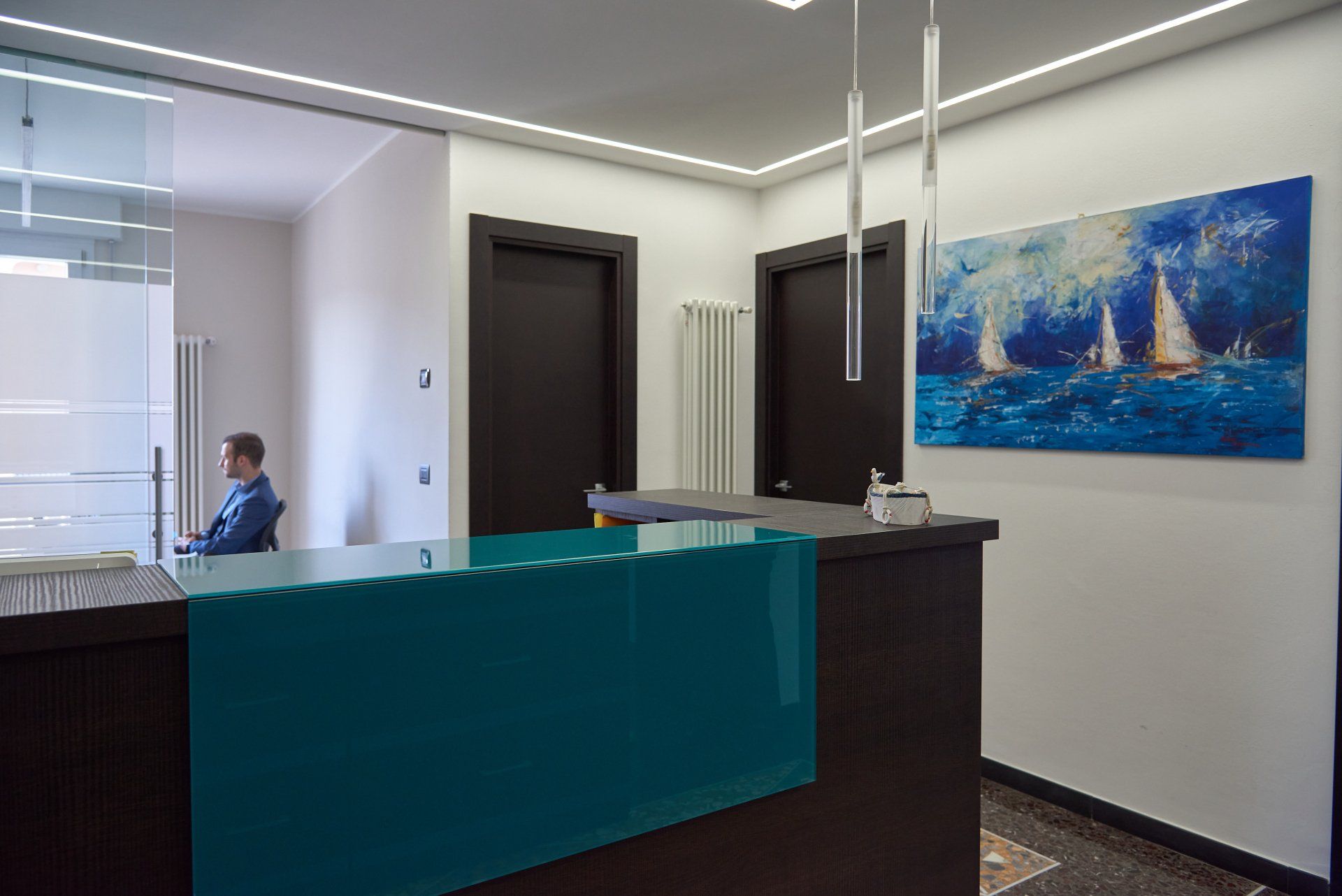 Area reception con bancone turchese, dipinto di barche a vela e una persona seduta sullo sfondo.