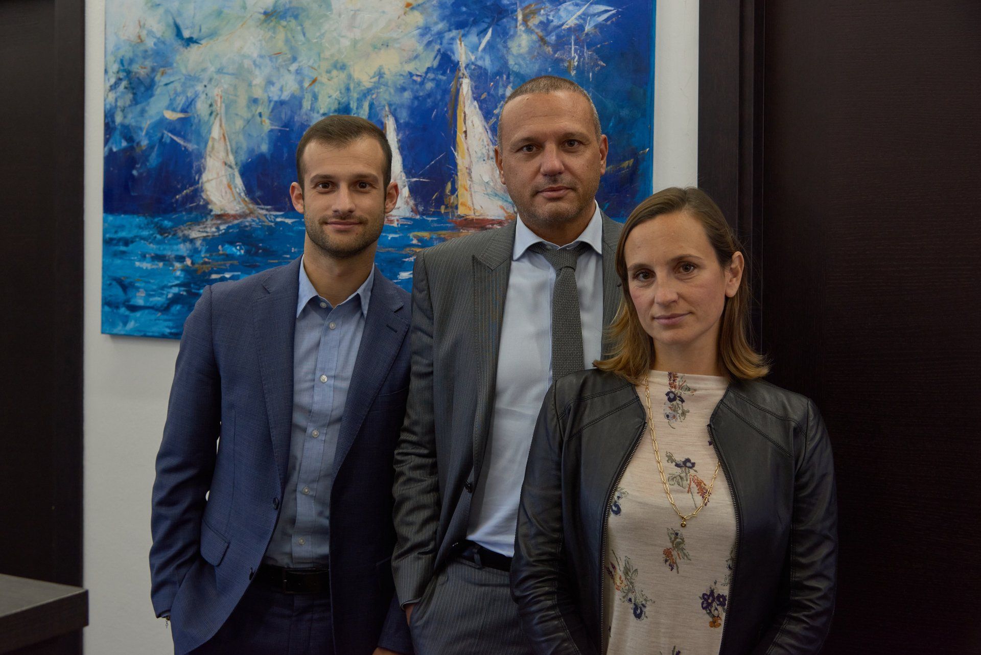 Tre professionisti posano davanti a un dipinto raffigurante una barca a vela blu. Due uomini e una donna, tutti in abiti da lavoro.