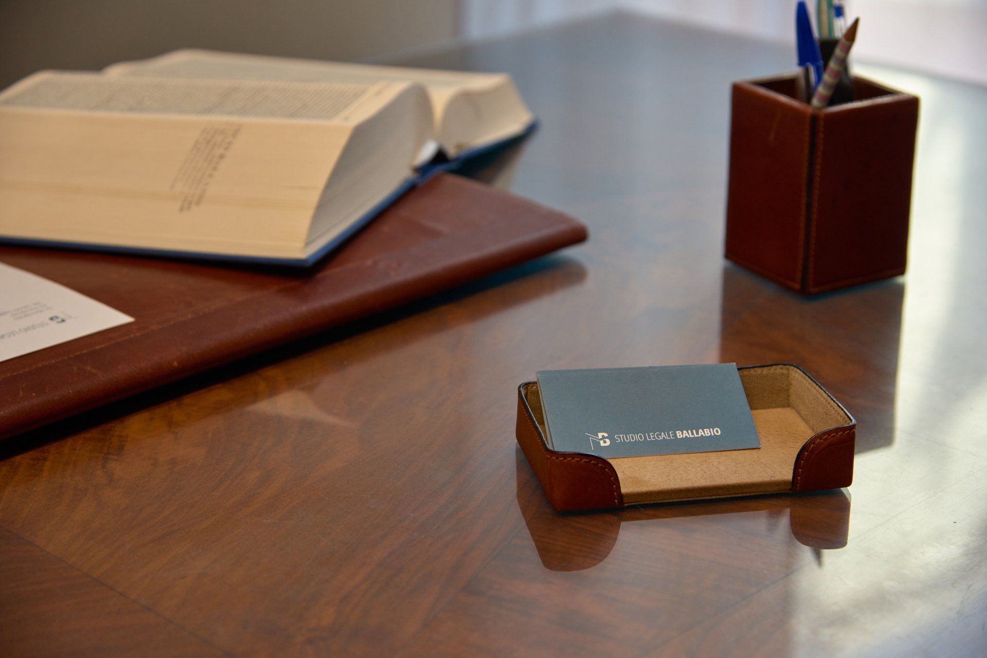 Scrivania con libro aperto, blocco note in pelle, portapenne e porta biglietti da visita.