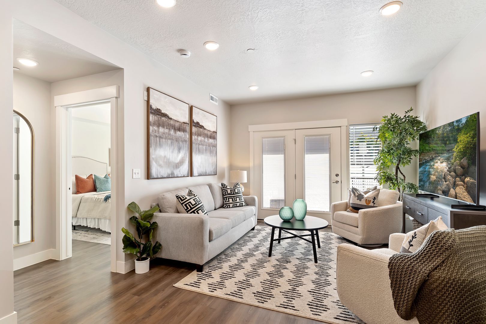 Living room with couch, rug, art, and TV, leading to a bedroom and French doors.