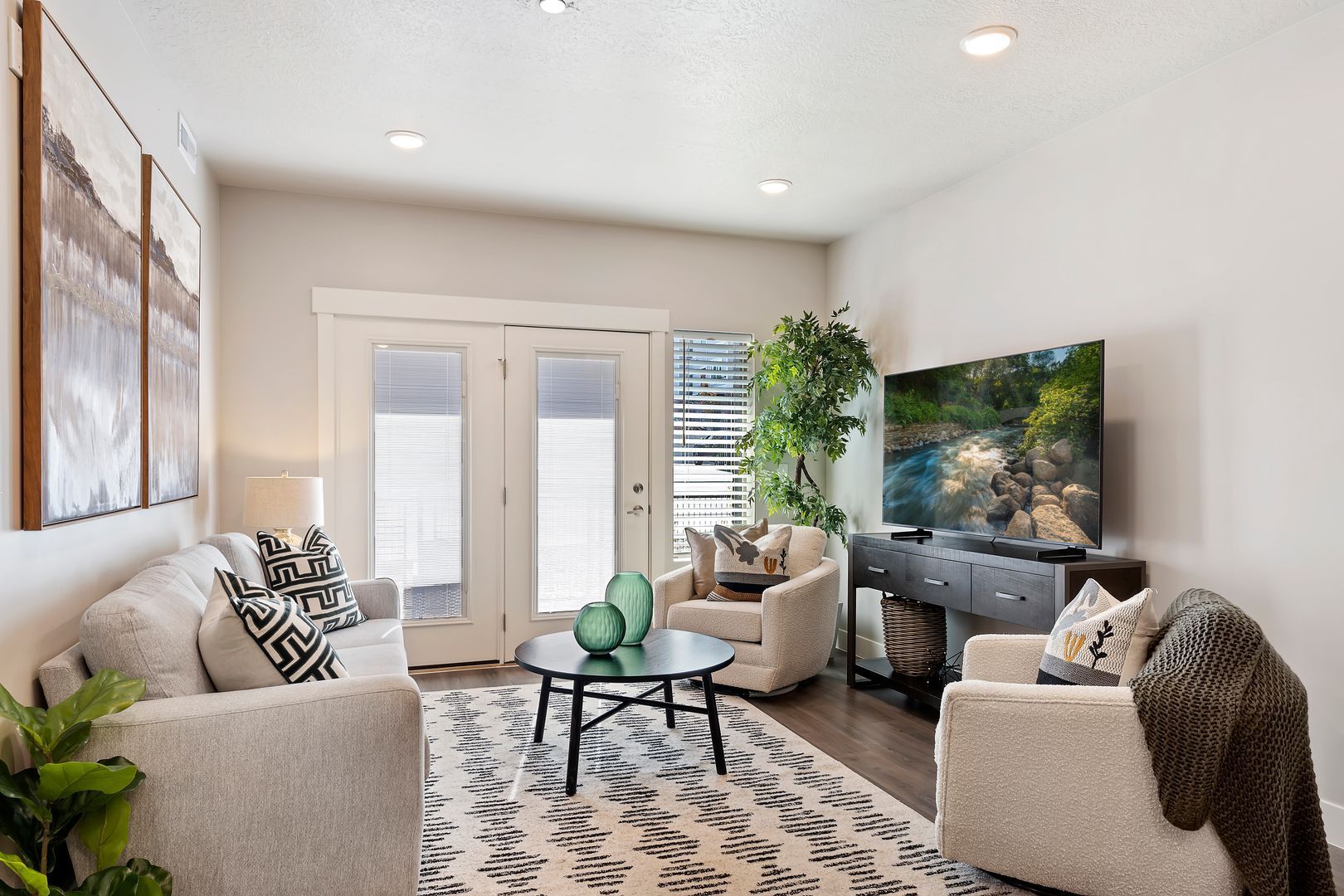 Living room with cream-colored sofas, patterned rug, TV, glass coffee table, and French doors leading outside.