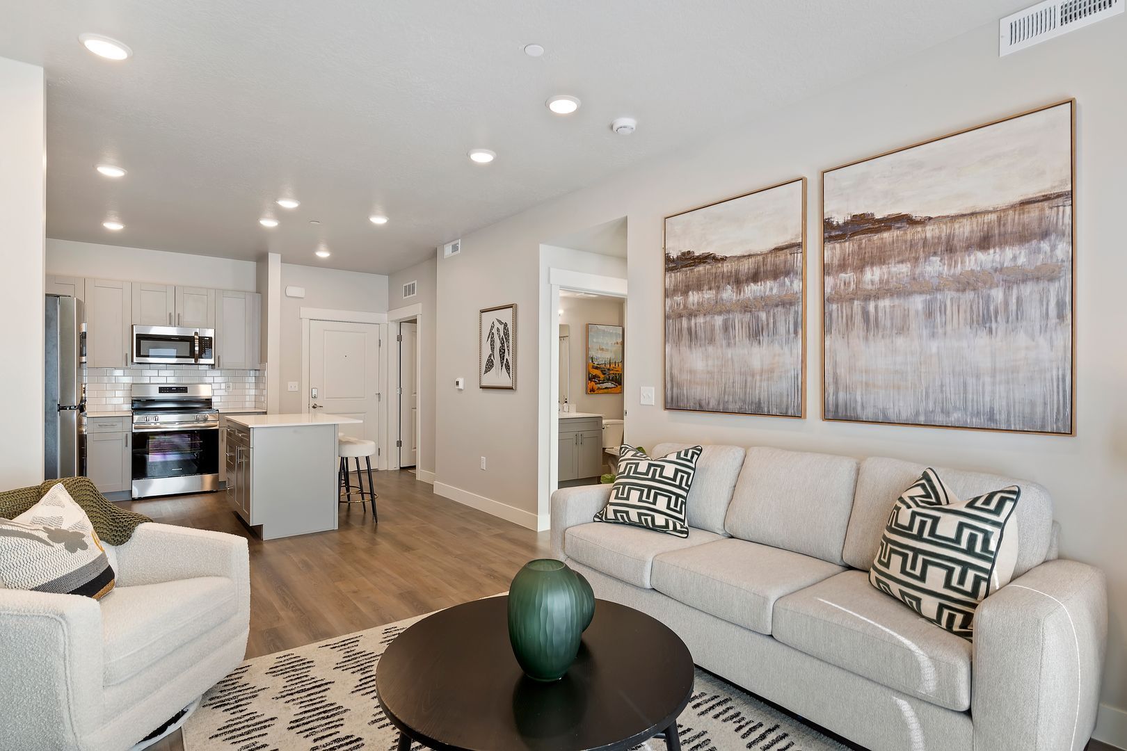 Open-concept living space with a kitchen, living area, and artwork, featuring light gray walls and wood flooring.