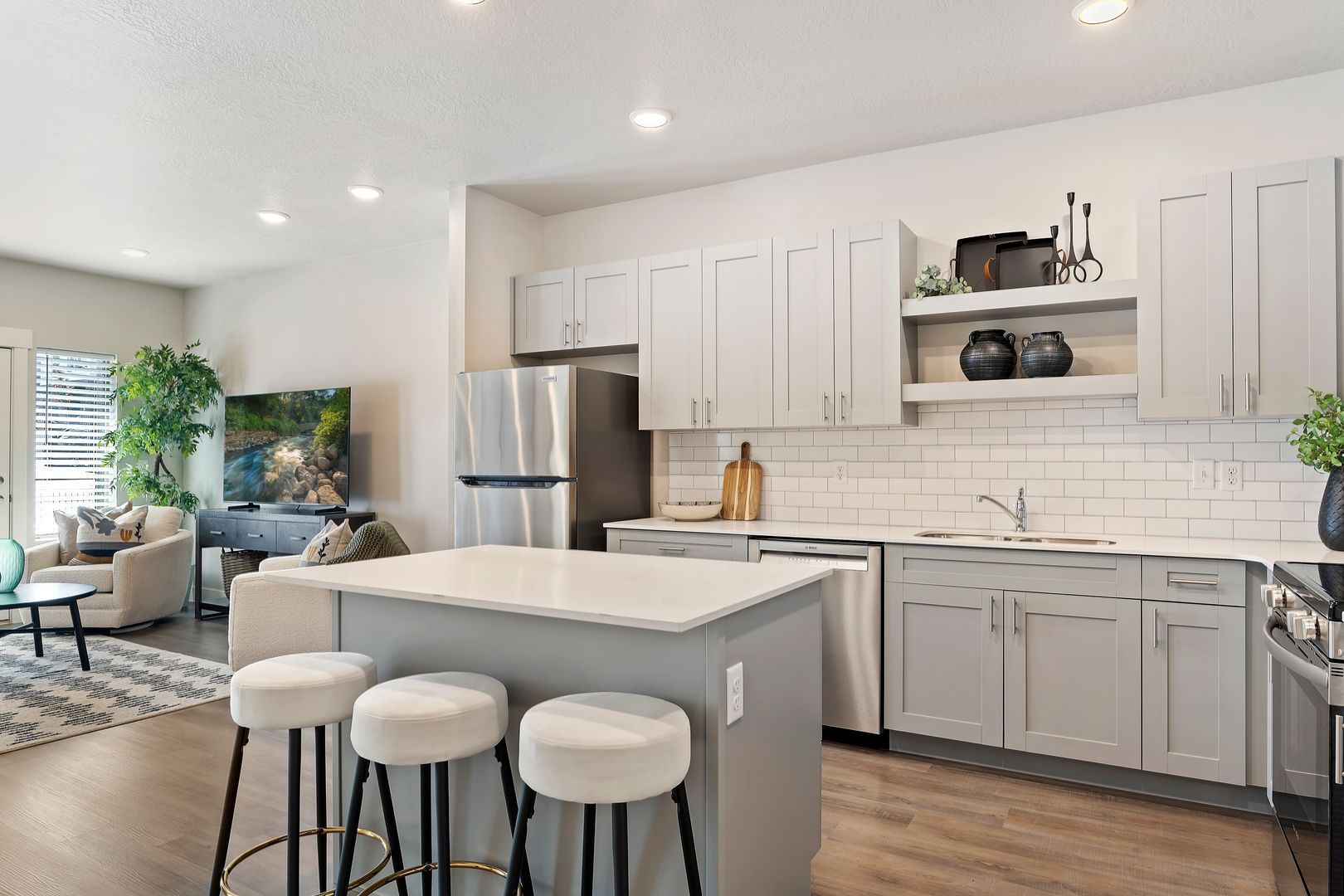 Modern kitchen with gray cabinets, stainless steel appliances, and island with stools; open to living area.