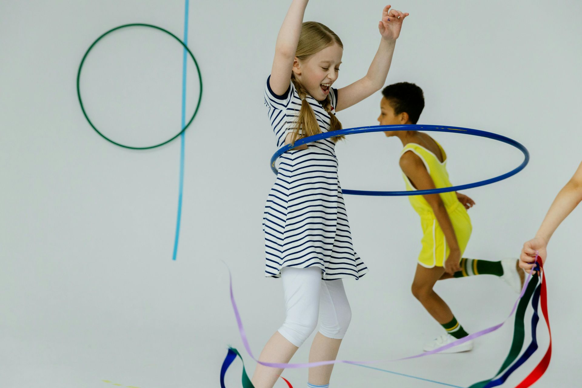 Girl jumping with hula hoop, another child runs, colorful ribbons and hoops against a white background.