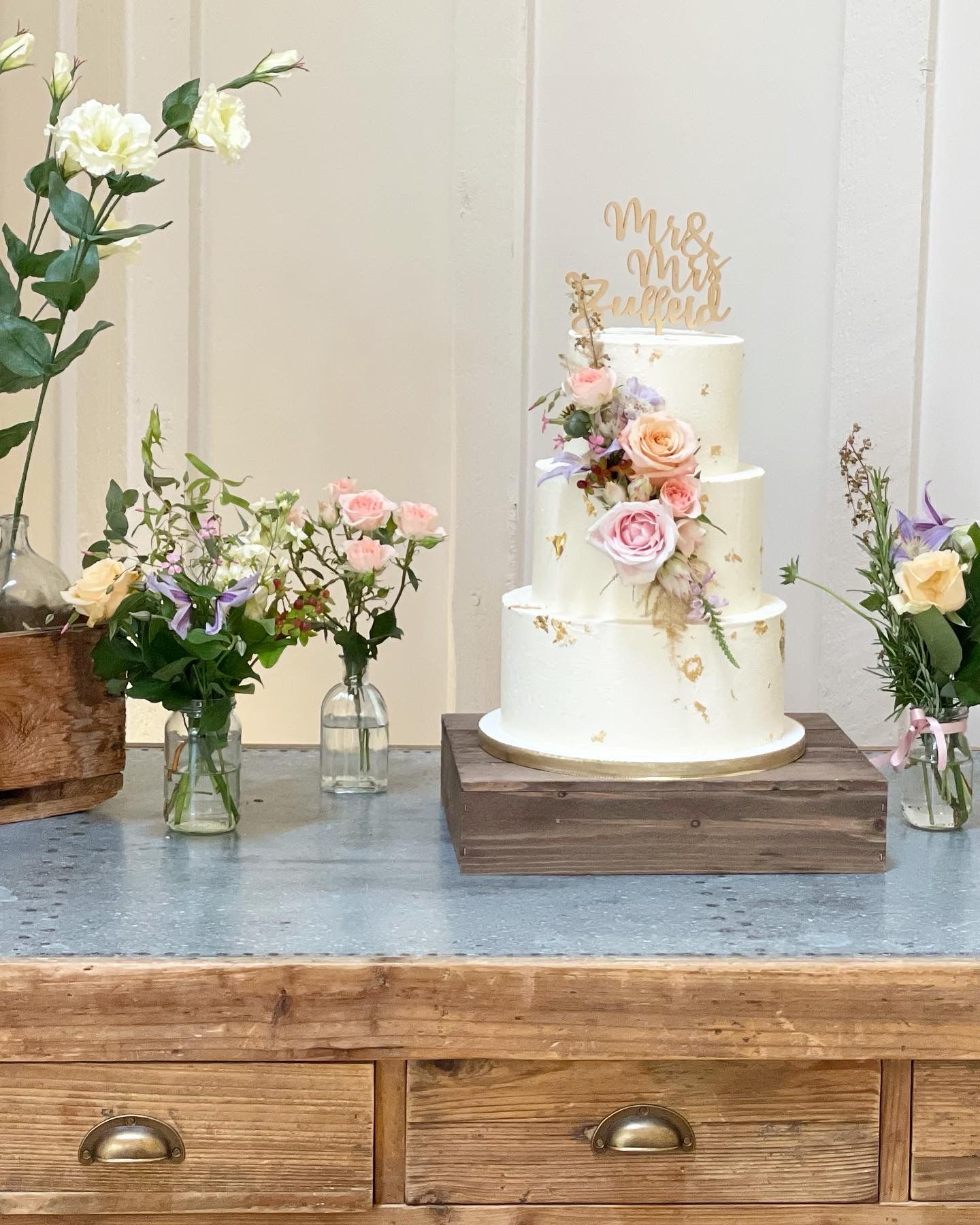 A wedding cake is sitting on top of a wooden table next to vases of flowers.