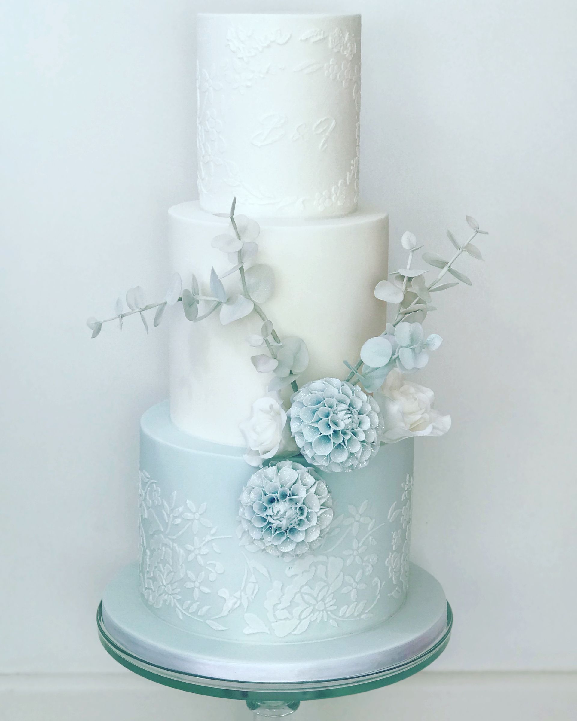 A white wedding cake with blue flowers is sitting on top of a glass cake stand.