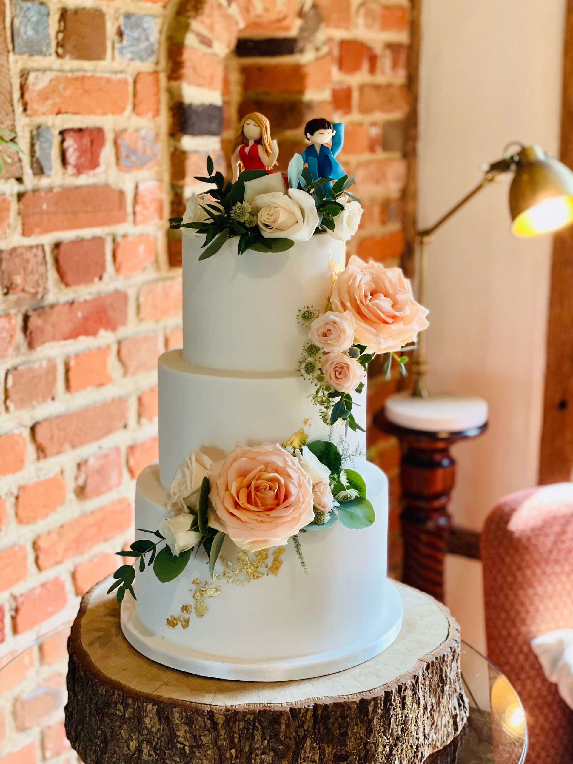 A wedding cake is sitting on a wooden stump in front of a brick wall.