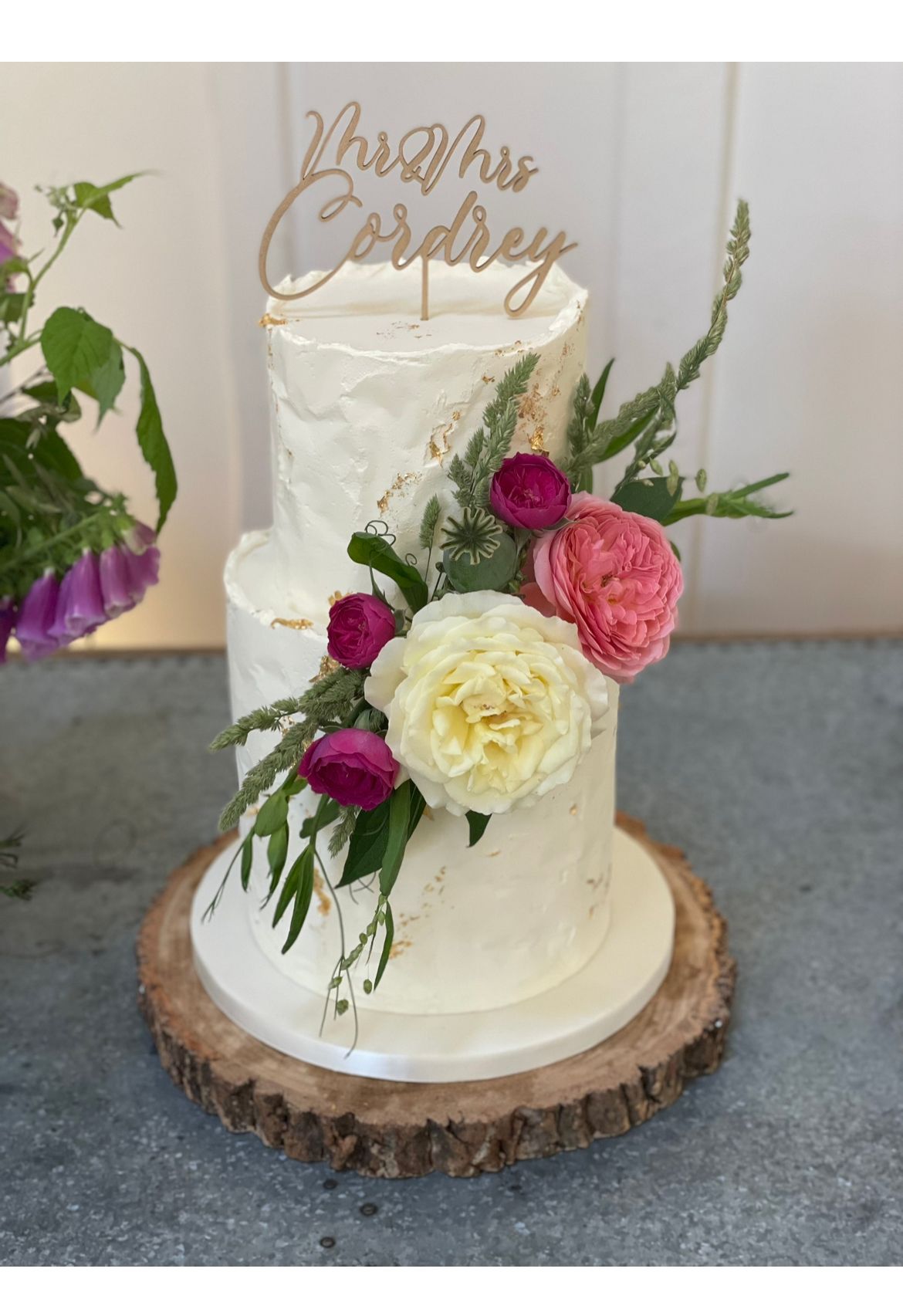 A wedding cake with flowers and a cake topper on a wooden slice.