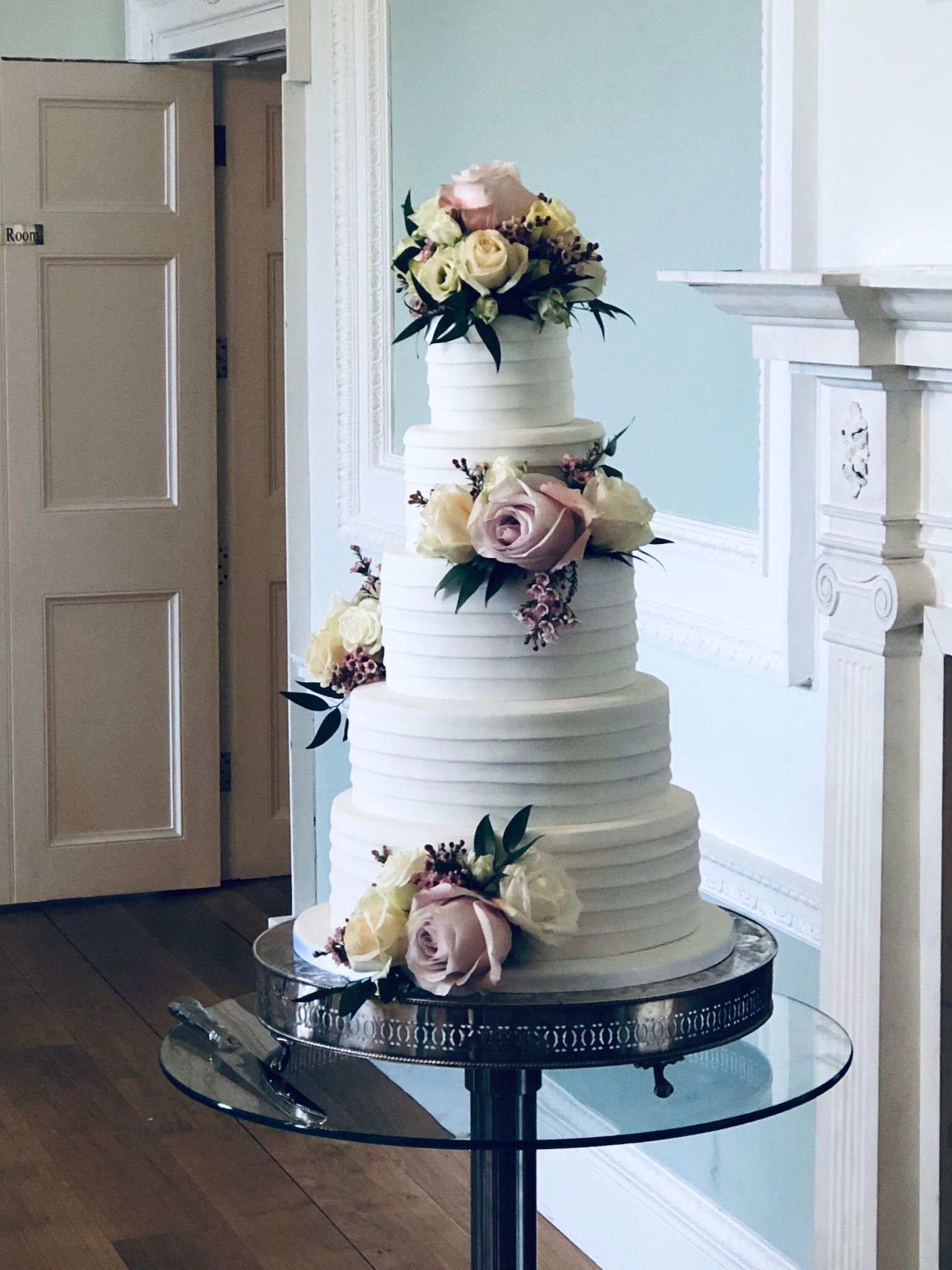 A wedding cake is sitting on a glass table in front of a fireplace.