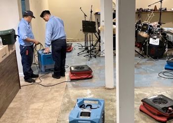 Two men are working on a machine in a room with drums.