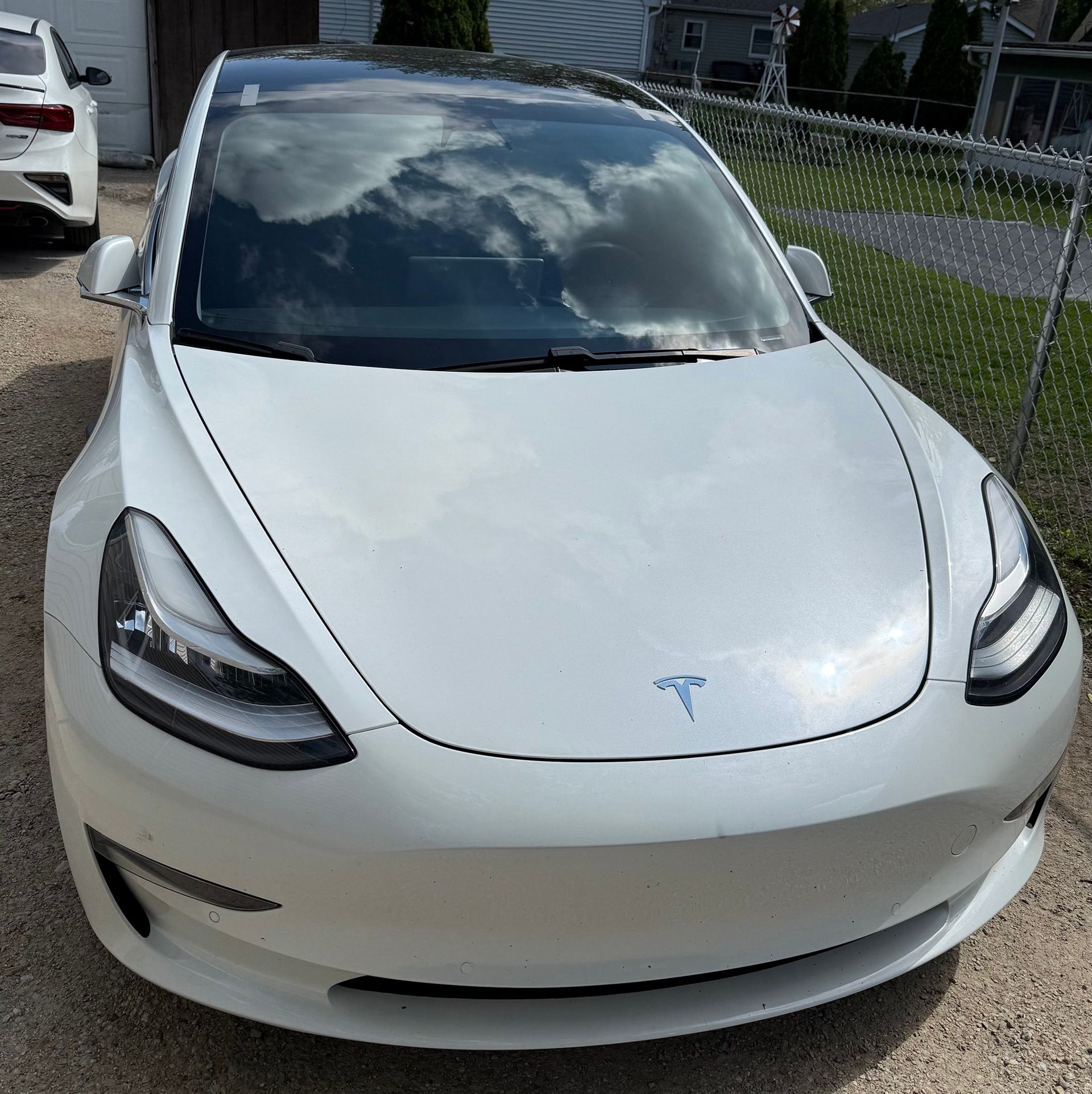 A white tesla model 3 is parked in a driveway next to a fence.