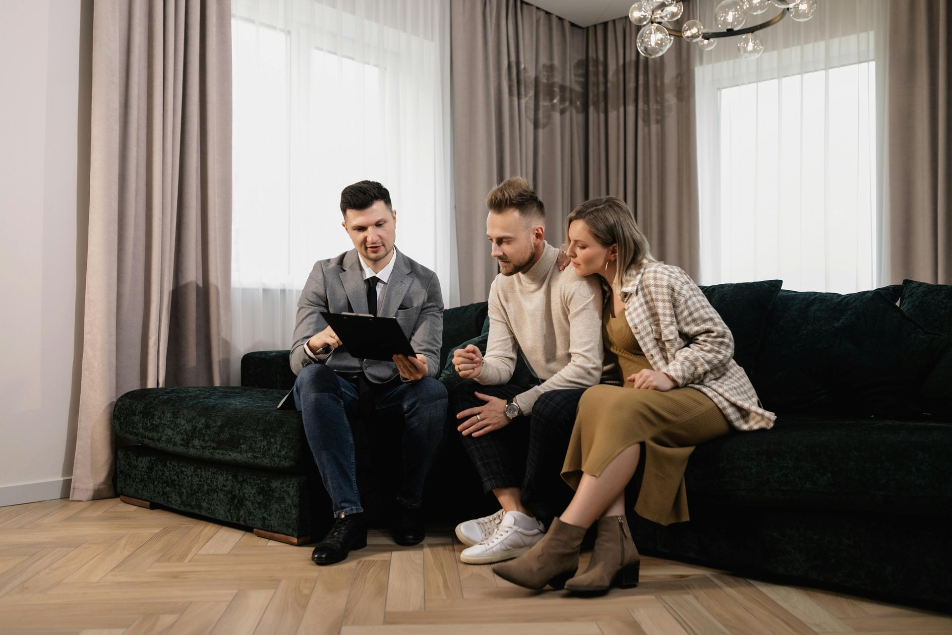 Three people sit on a dark green sofa, looking at a tablet in a bright living room.