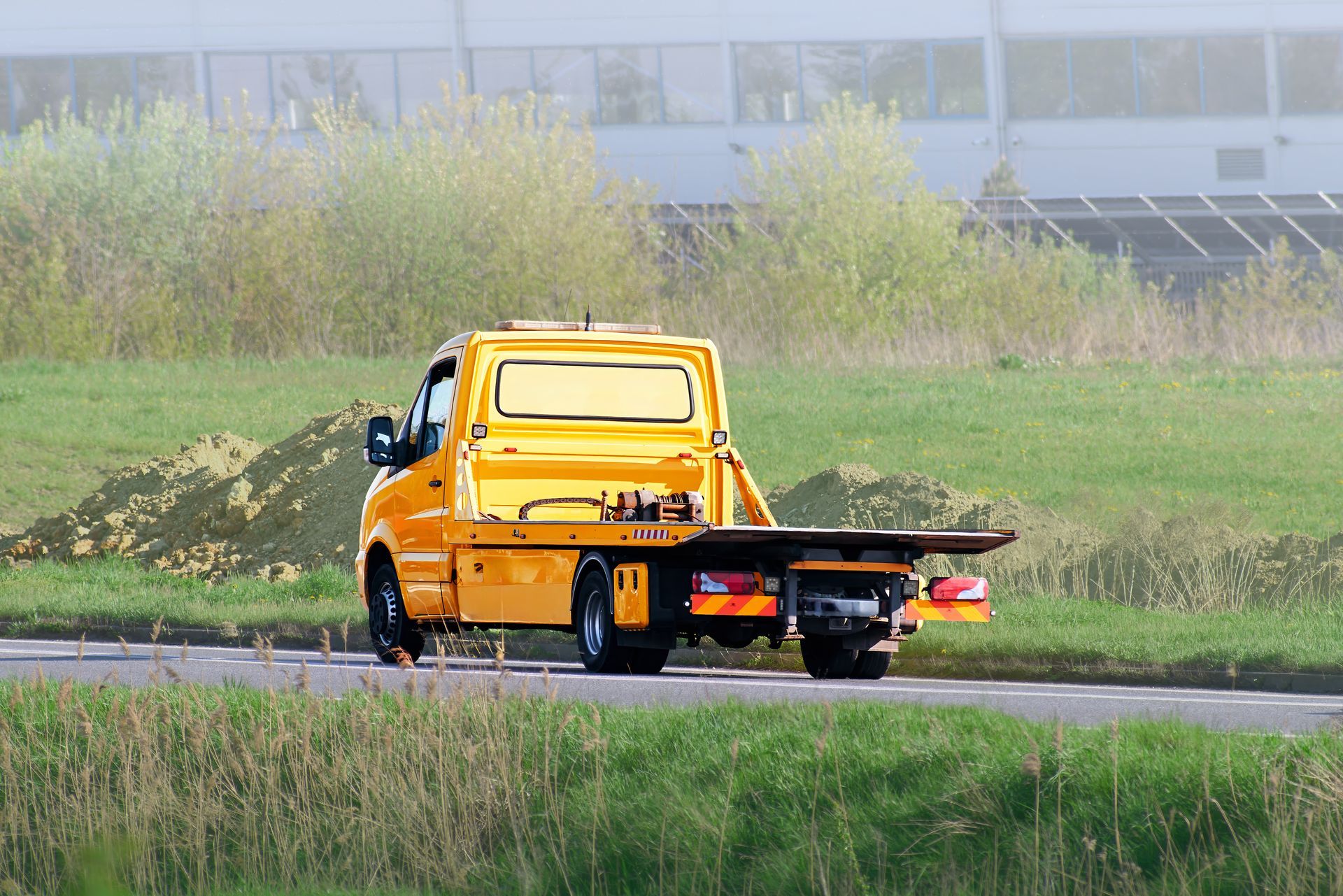 Yellow tow truck on a road, transporting vehicles. Green fields, building in background.