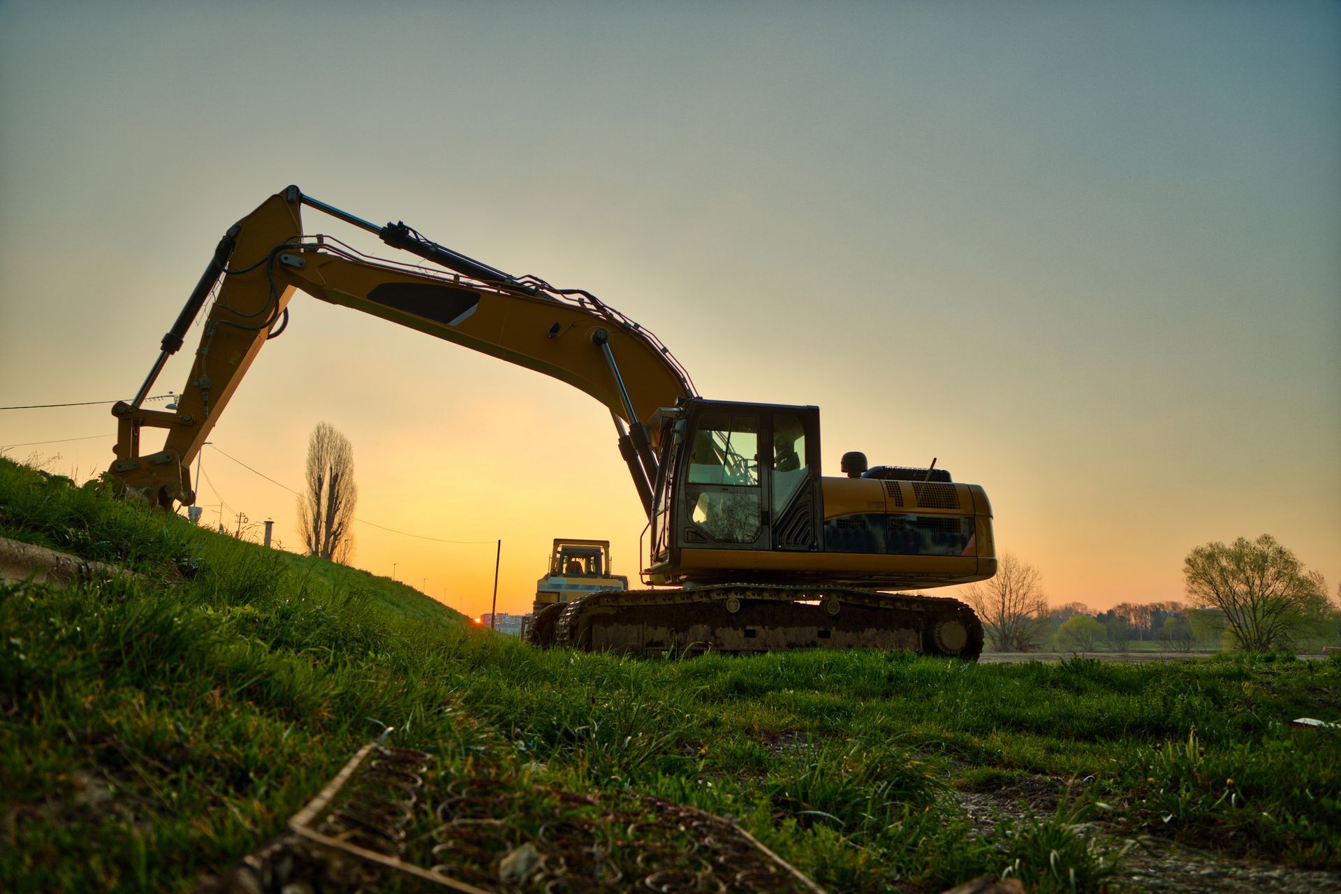 Yellow excavator on a grassy hill with a sunset background.