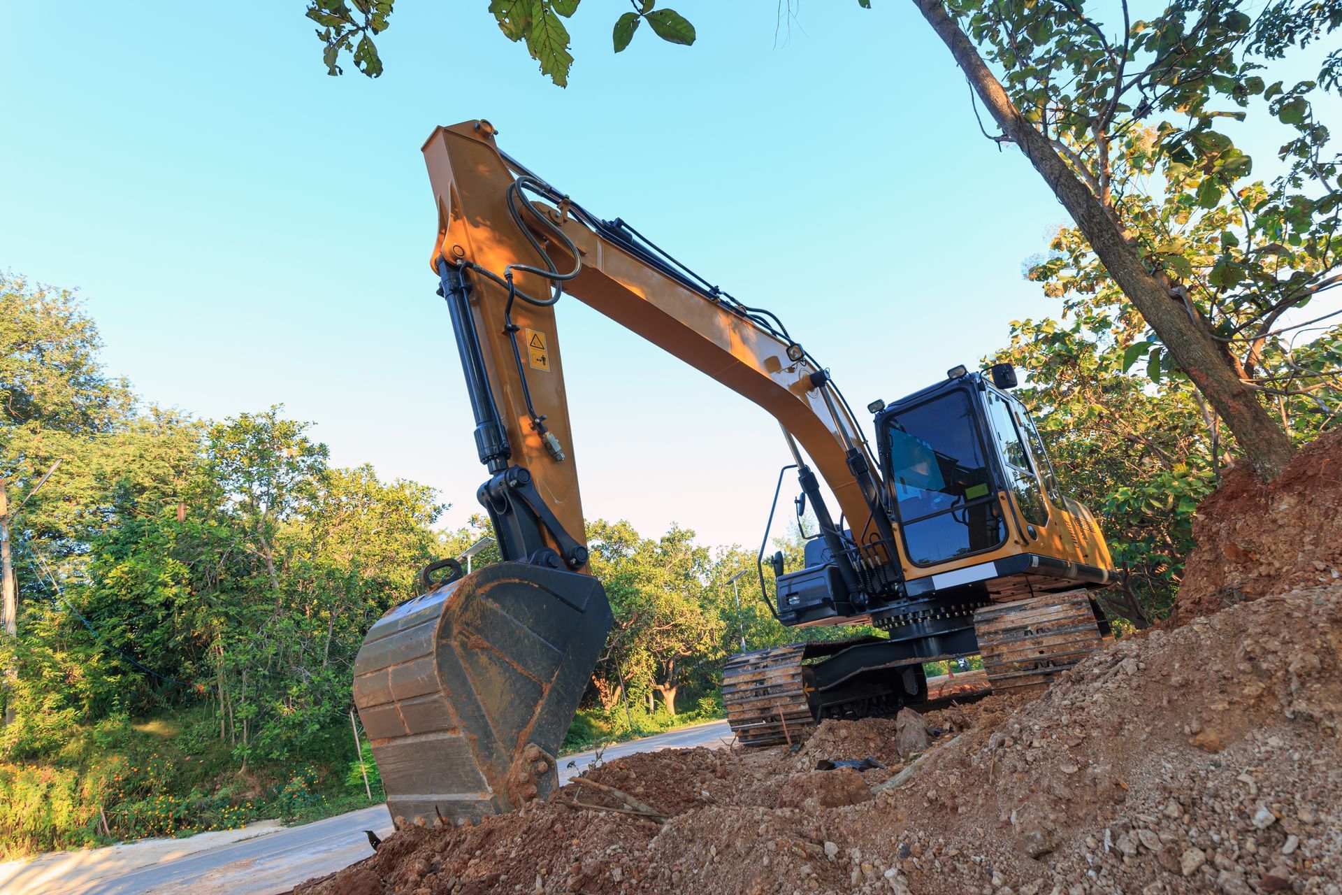 Yellow excavator digging on a hillside, trees in the background, clear sky.