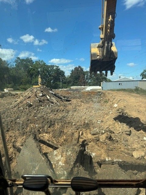 Excavator demolishing concrete and earth on a construction site; blue sky, trees in the background.