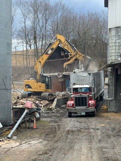 Excavator demolishing building; semi-truck parked nearby. Debris scattered. Cloudy day.