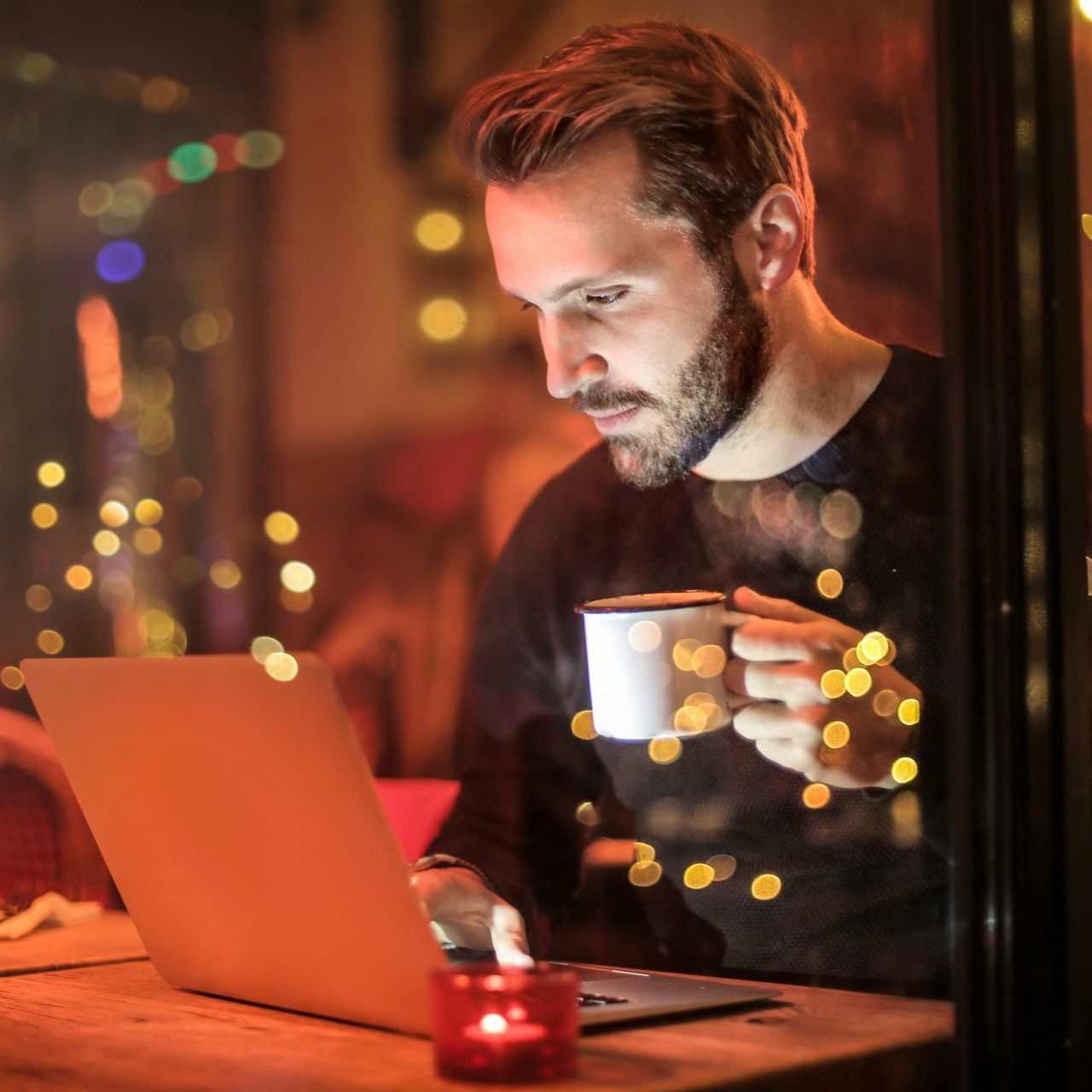 Man at a laptop, holding a mug, in a warm-lit room with blurred lights in the background.