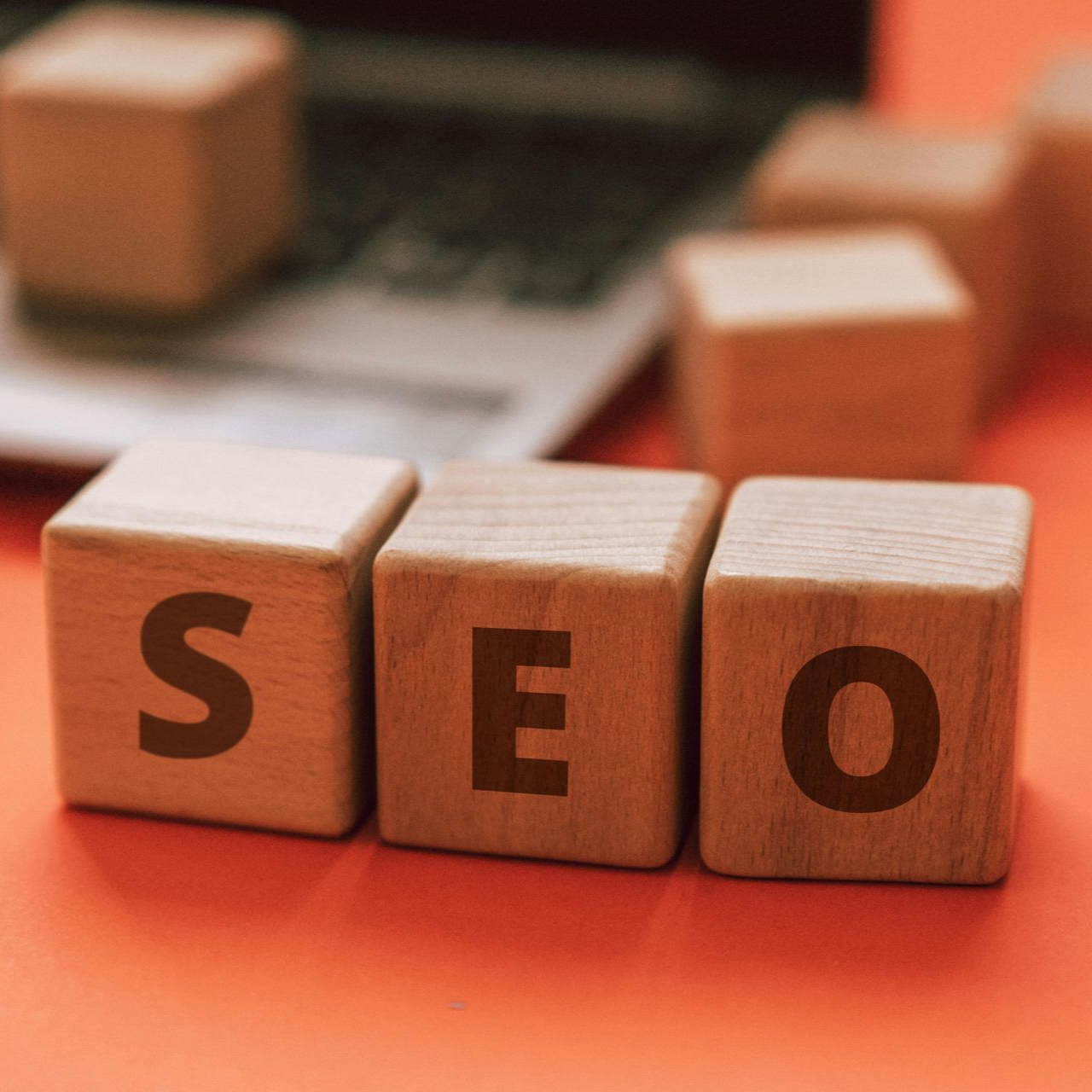 Wooden blocks spelling SEO on orange surface, with blurred laptop in background.
