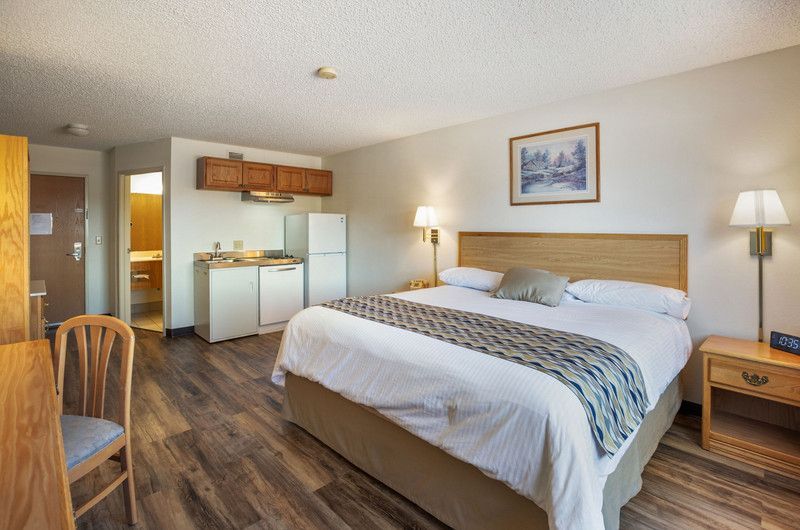 A hotel room with a king size bed , desk , chair and refrigerator, kitchenette and window.