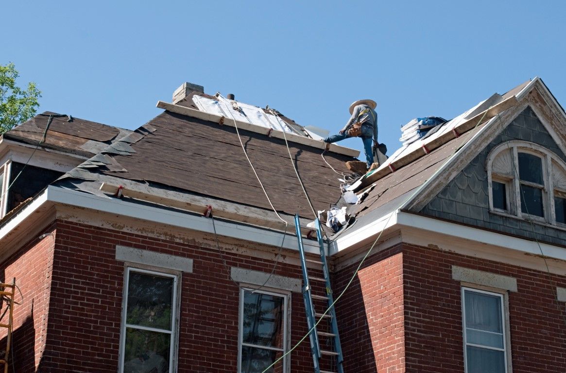 Roofer on a ladder replacing shingles on a brick house under a blue sky.