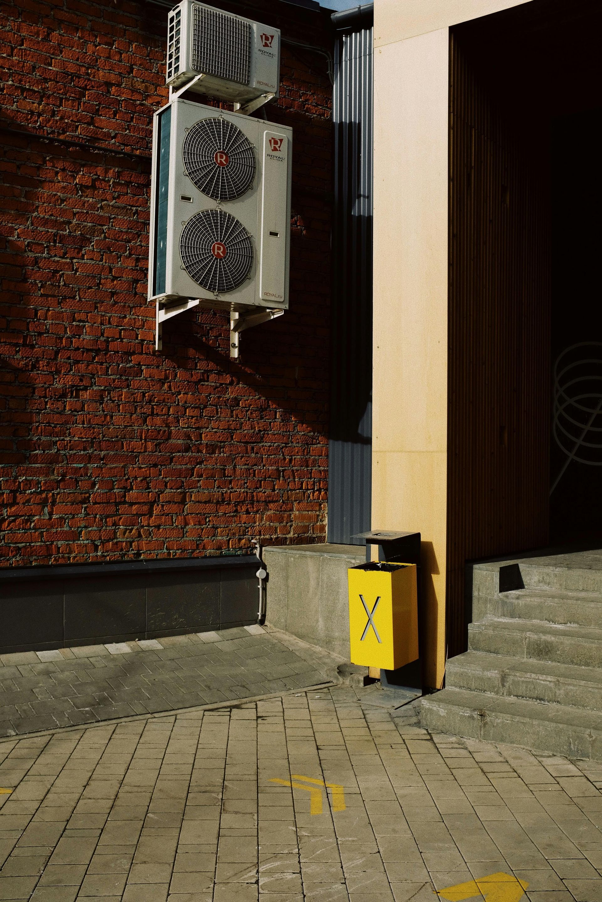 Brick wall with air conditioning units and a yellow trash can next to a door.
