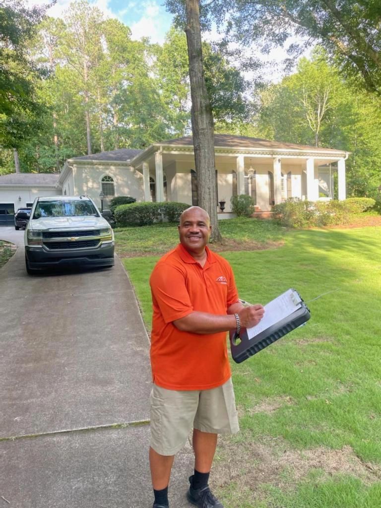 A man in an orange shirt is standing in front of a house holding a clipboard.