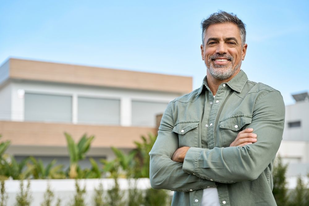 A man is standing in front of a house with his arms crossed.
