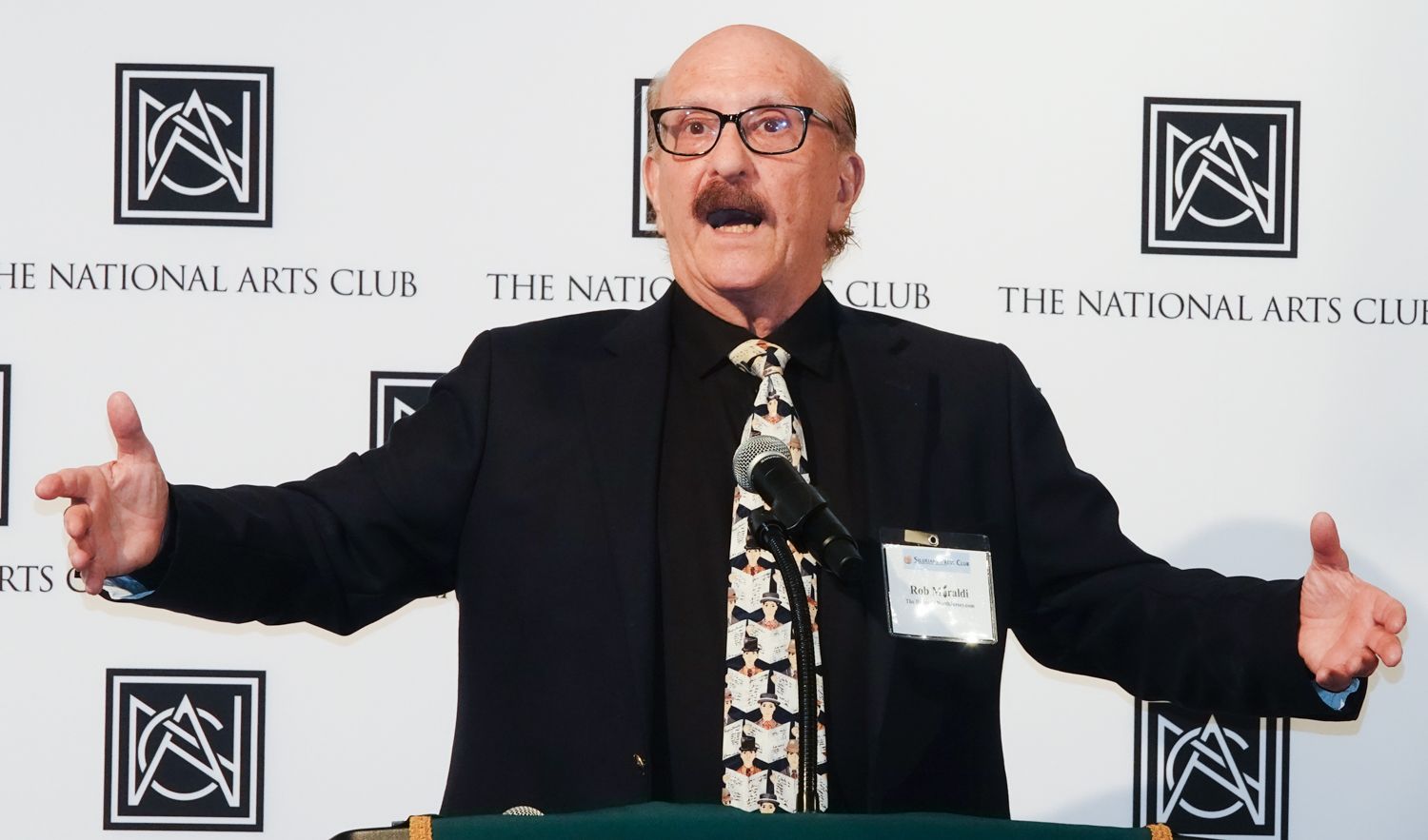 A man stands at a podium in front of a wall that says the national arts club