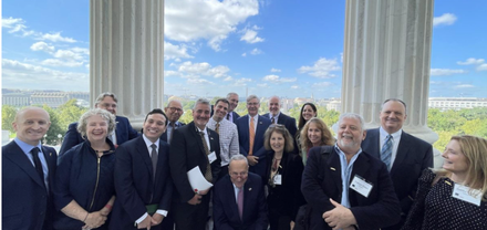Group of people posing outdoors with a building in the background. Many are wearing suits and smiling. Sunny day.