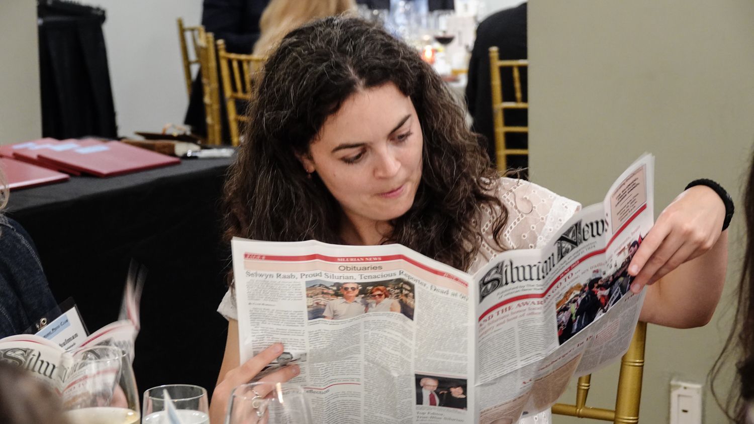 A woman is reading a newspaper while sitting at a table.