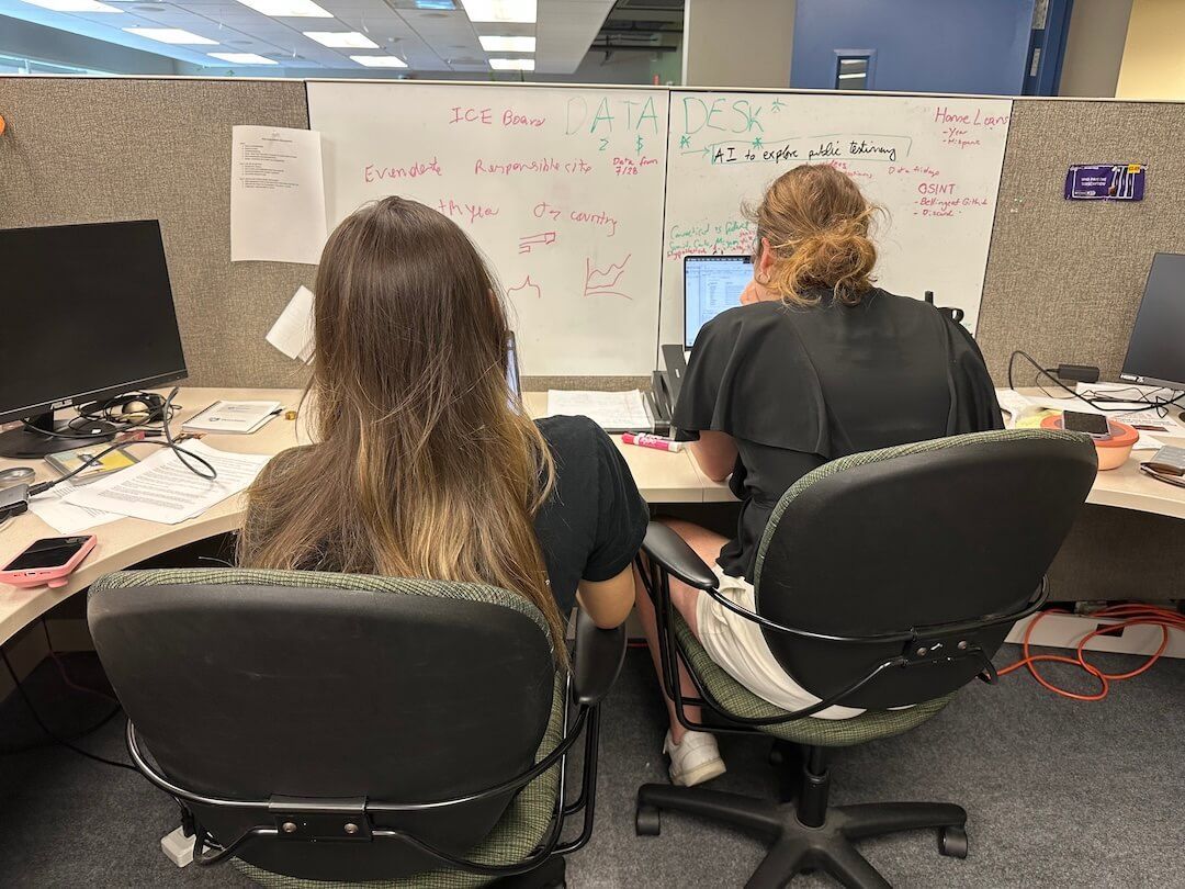 Two people at adjacent office cubicles, working at their desks with whiteboards behind them.