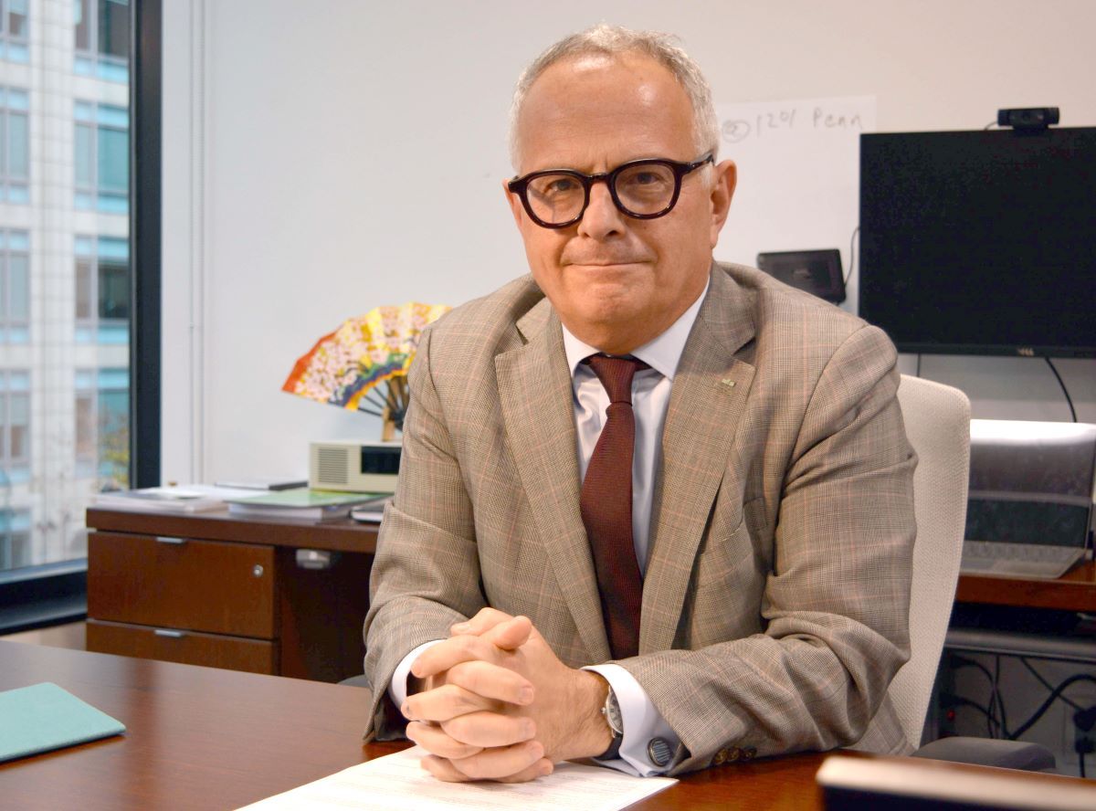 Man in glasses and suit, sitting at a desk, hands clasped. Office setting, natural light.