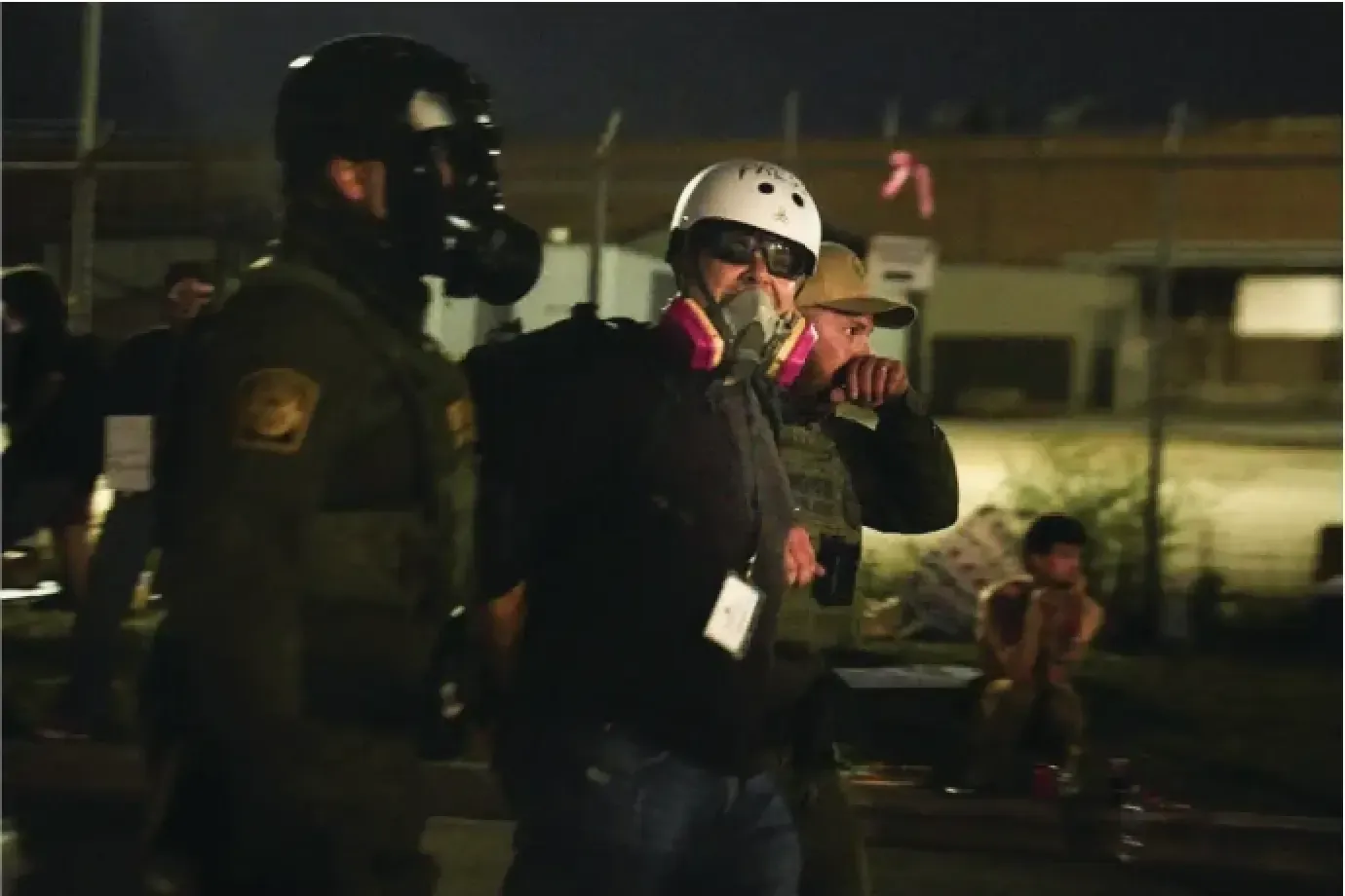Two figures in protective gear stand near a chain-link fence at night. A child is visible in the background.