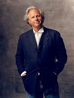 A person with gray hair wearing a dark blazer and white shirt stands with hands in their pockets against a gray backdrop.