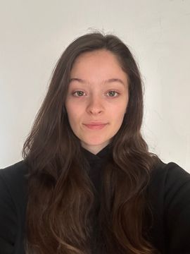 Young person with long dark hair in a black top, facing the camera against a plain light wall.