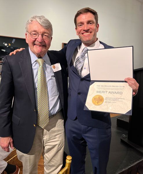 Two men pose at an event; one holds a framed award. Man on left wears a blazer and tie; man on right wears a suit and tie.