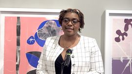 Woman speaking at a podium, wearing glasses and a blazer, in front of abstract art on a wall.