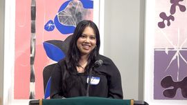 Woman speaking at a lectern with artwork in background. She smiles with a nametag on, in a gallery setting.