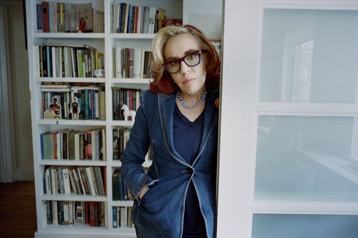 Woman in glasses, denim jacket, leaning in doorway by bookshelf.