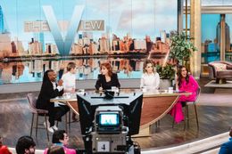 Talk show panel in a studio with host desk, camera, and city skyline backdrop