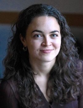 Woman with curly brown hair smiles, wearing a dark top, indoors.