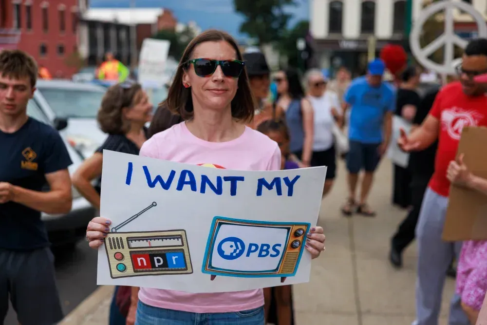A person in a pink shirt holds a sign saying "I WANT MY" with illustrations of an NPR radio and a PBS television.