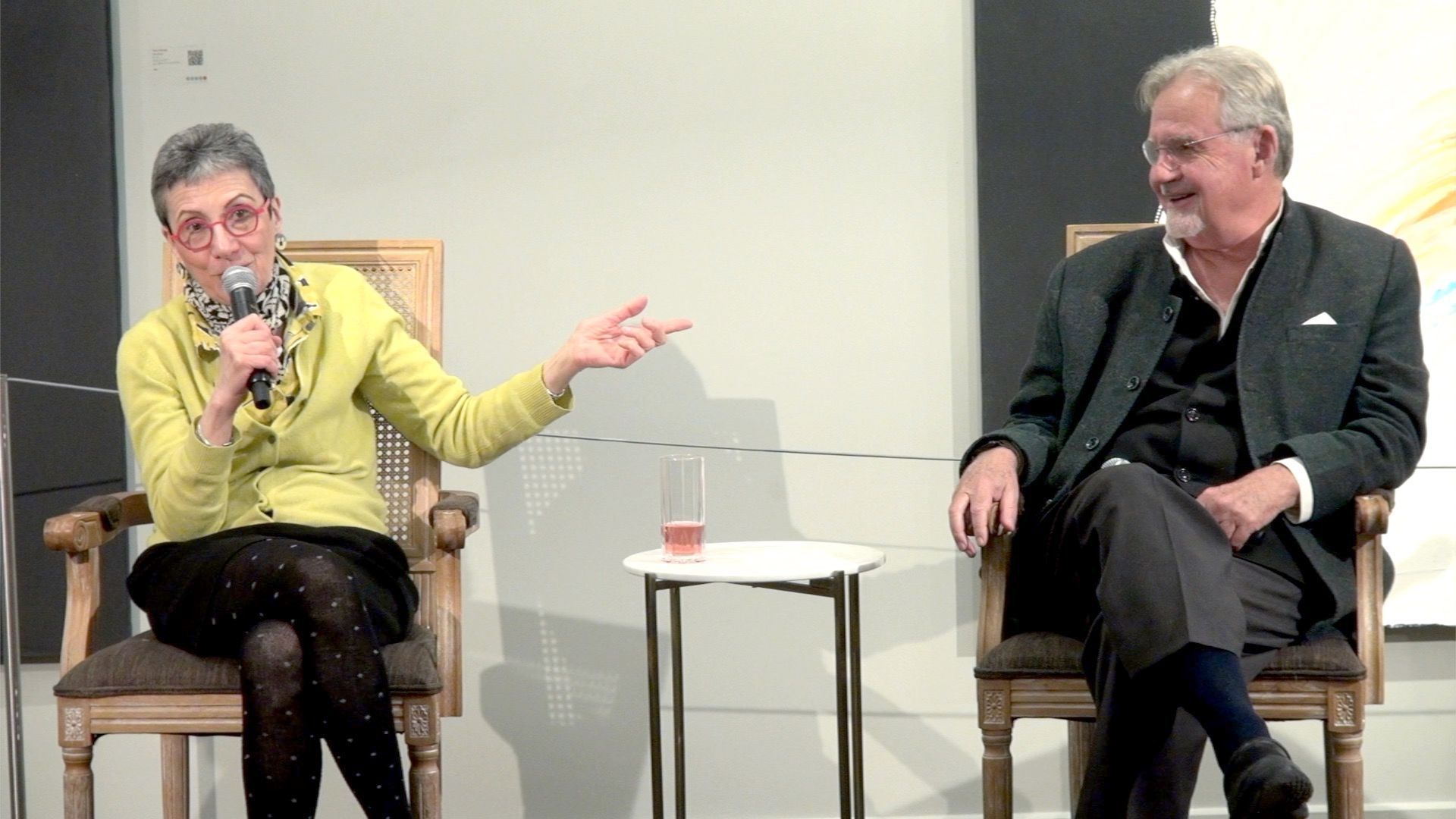 Two people sit in chairs on a stage during a presentation, with a speaker holding a microphone and gesturing to the side.