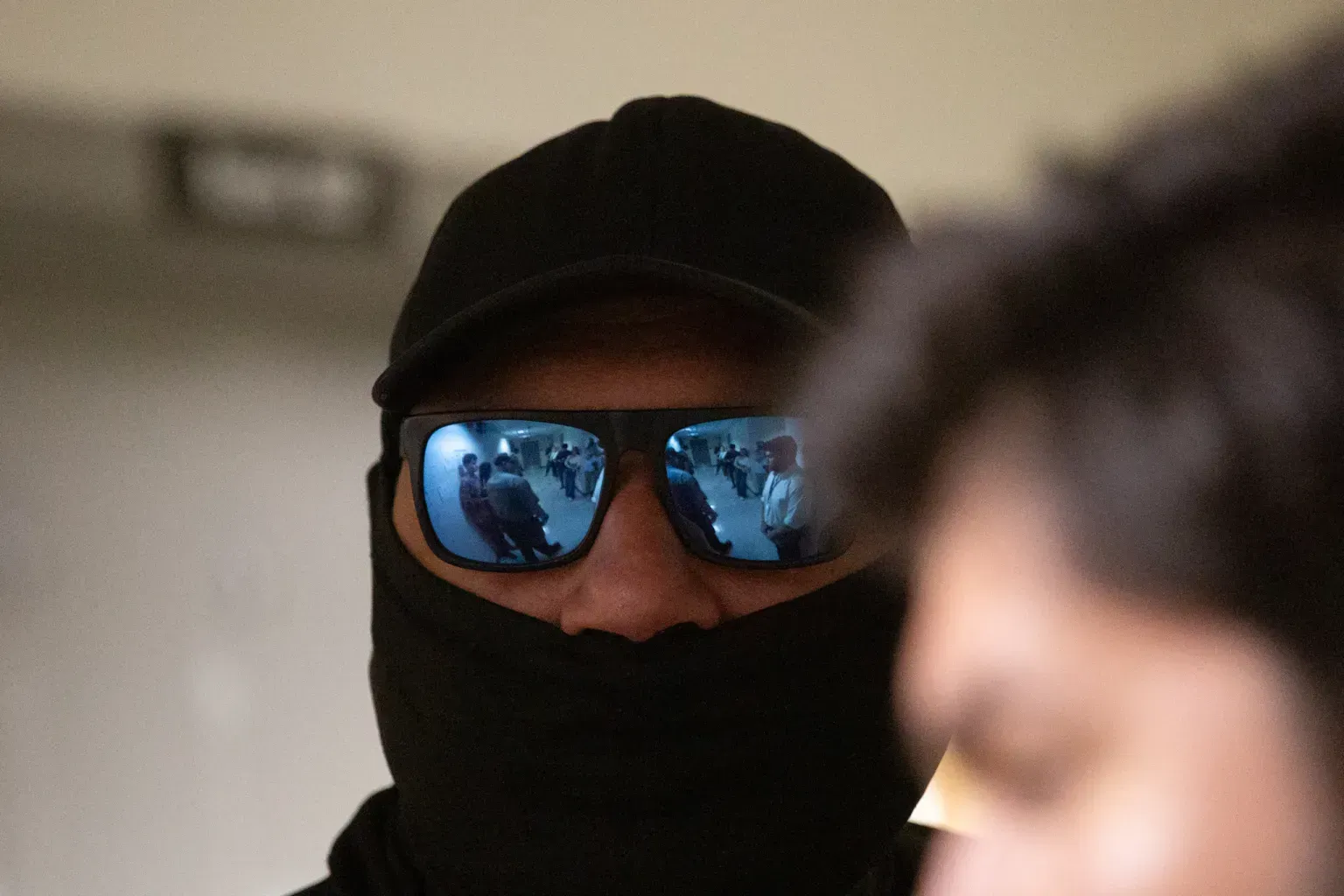 Masked person wearing blue reflective sunglasses and a black cap, with a blurred person in the foreground