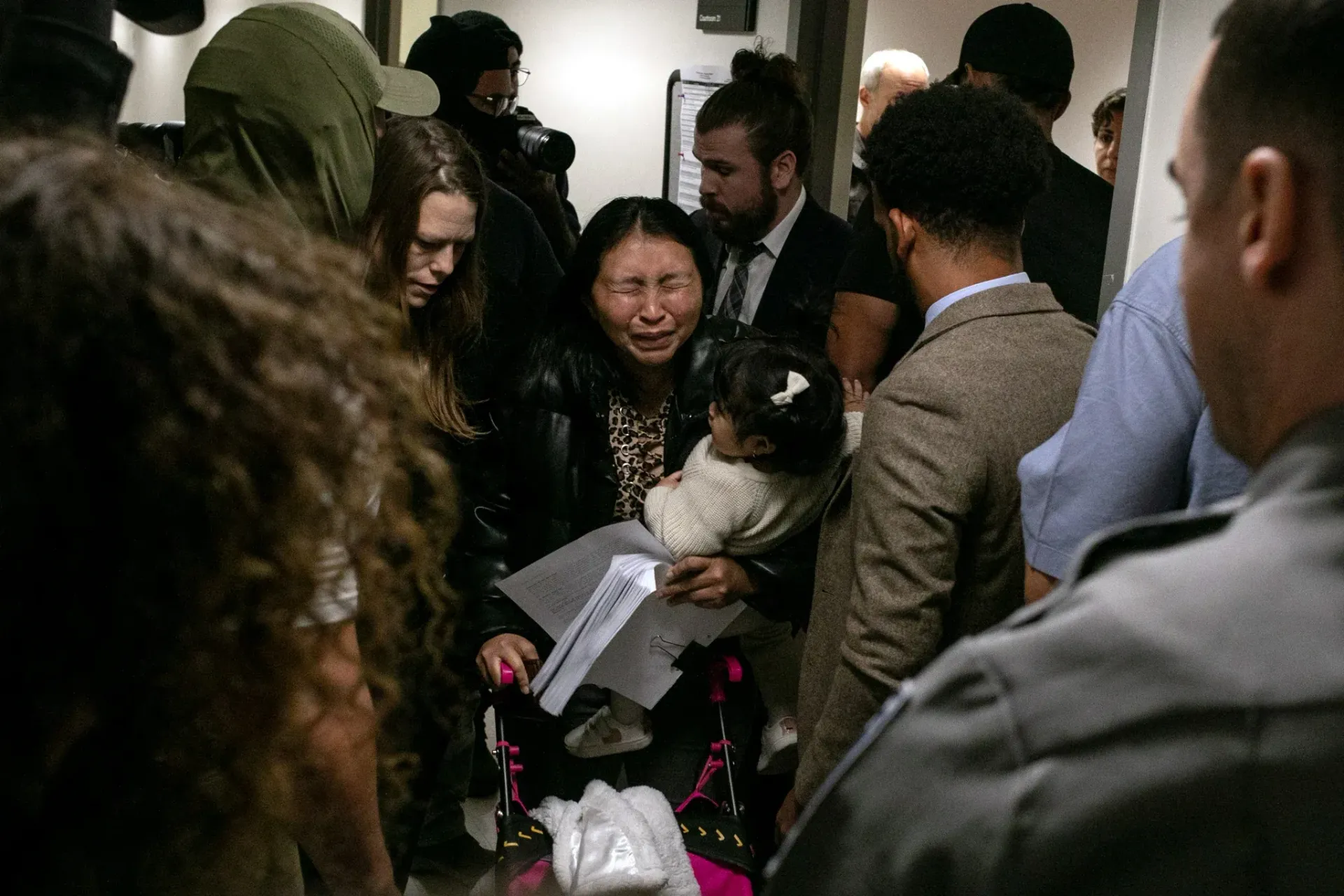 Crowded indoor scene with people gathered around a seated person holding papers and a bouquet