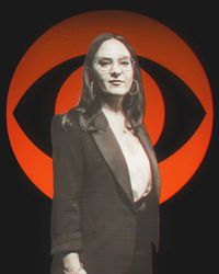 Person in a dark blazer stands before a large orange eye-like logo on a black background.