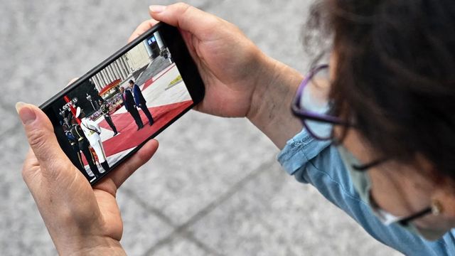 Person holding a smartphone horizontally, watching a formal event on the screen.